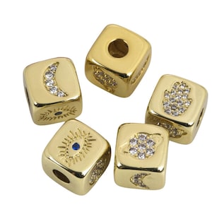 Op de afbeelding: Vijf vierkante kralen in goudkleur met verschillende symbolen en heldere strasssteentjes. De symbolen omvatten een halve maan, een hamsa-hand, een boze oog, een planeet en een cirkel met een lijn erdoorheen.