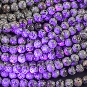 May include: A close-up of a strand of round, speckled, gray and white beads. The beads are illuminated with a bright purple light, highlighting the intricate patterns within each bead.