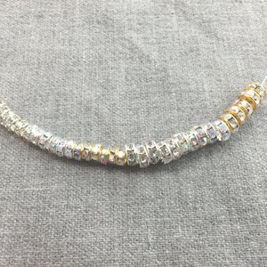 May include: A string of cylindrical beads threaded on a thin wire. The beads alternate between silver-toned metal with clear, iridescent rhinestones and gold-toned metal bands. The beads are arranged in a curved line, showcasing a mix of metallic finishes and sparkling, light-reflecting crystal accents throughout the entire length of the strand.