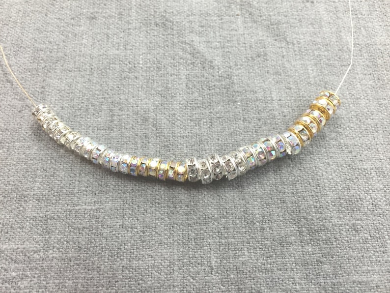 May include: A string of cylindrical beads threaded on a thin wire. The beads alternate between silver-toned metal with clear, iridescent rhinestones and gold-toned metal bands. The beads are arranged in a curved line, showcasing a mix of metallic finishes and sparkling, light-reflecting crystal accents throughout the entire length of the strand.