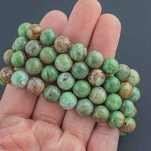 May include: A strand of green and brown gemstone beads, likely variscite, in a hand. The beads are round and have a mottled appearance.
