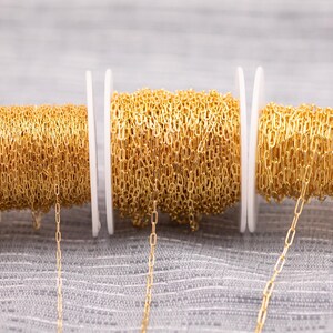 May include: Three spools of gold-colored chain are displayed against a gray background. The chain is made of small, rectangular links. The spools are white and hold the chain in neat coils. A few strands of chain hang down from the spools.