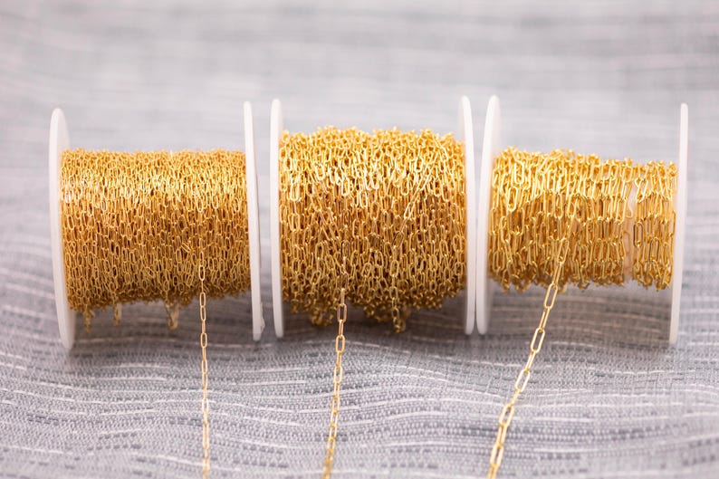 May include: Three spools of gold-colored chain are displayed against a gray background. The chain is made of small, rectangular links. The spools are white and hold the chain in neat coils. A few strands of chain hang down from the spools.