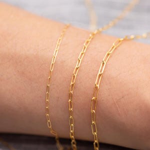 May include: Close-up of three delicate gold-colored paperclip chain necklaces. The chains are thin and feature a repeating rectangular link design. The necklaces are displayed against a neutral background, highlighting their bright, shiny appearance. Jewelry.