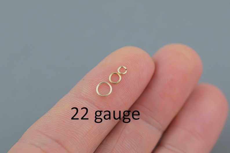 May include: Three small, gold-colored jump rings of varying sizes are displayed on a fingertip against a gray background. The text "22 gauge" is printed in black below the rings, indicating the wire thickness.