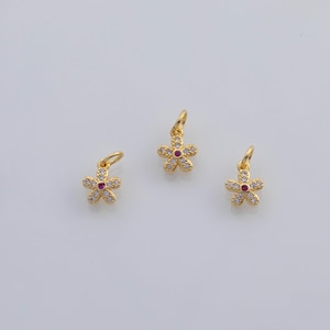 Op de afbeelding: Drie goudkleurige bloembedels. Elke bedel heeft een bloemontwerp met vijf bloemblaadjes, een rood centrum en heldere kristallen accenten. De bedels hebben een kleine lus aan de bovenkant om aan een ketting of armband te bevestigen.