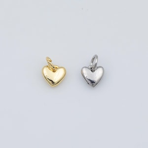 May include: Two heart-shaped pendants. One is gold-coloured, the other is silver-coloured. Each pendant has a small loop at the top for attaching to a chain or bracelet. The pendants are displayed on a white background.