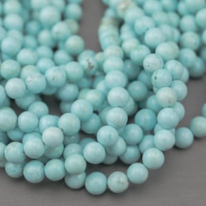 May include: Close-up of strands of light blue round beads. The beads have a matte finish and are strung together, creating a flowing, organic shape. The beads are a consistent size and color, with subtle variations.