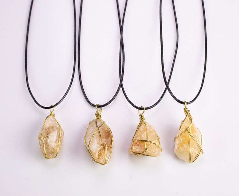 May include: Four necklaces displaying raw, golden-yellow citrine crystals, each encased in gold-coloured wire. The crystals hang from black cords. The crystals' shapes and sizes differ, highlighting their natural, unpolished form.