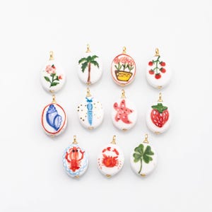 May include: A collection of twelve white oval pendants with gold-colored loops. Each pendant features a hand-painted design, including a rose, palm tree, flowers, strawberries, a seashell, a shrimp, a starfish, a strawberry, a lobster, a crab, and a four-leaf clover.