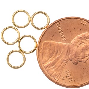 May include: Six small, gold-colored jump rings are arranged near a U.S. penny. The penny is dated 2021 and shows the profile of Abraham Lincoln. The jump rings are open and ready to be used for jewelry making or crafts.