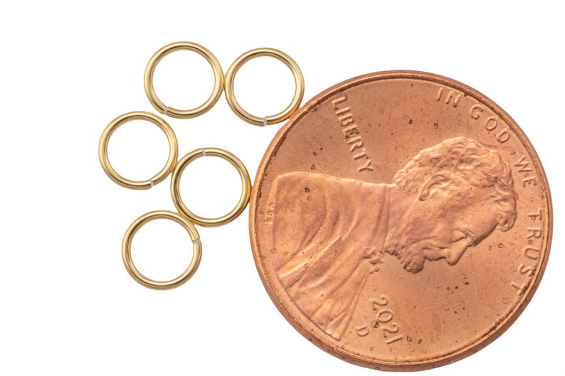 May include: Six small, gold-colored jump rings are arranged near a U.S. penny. The penny is dated 2021 and shows the profile of Abraham Lincoln. The jump rings are open and ready to be used for jewelry making or crafts.