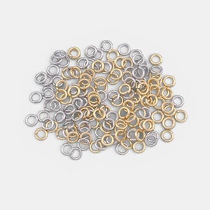 Puede incluir: Un conjunto de pequeños anillos abiertos en tonos dorados y plateados. Estos anillos metálicos están dispersos sobre una superficie blanca, ideales para la fabricación de joyas y proyectos de artesanía. Los anillos son circulares y tienen una abertura.