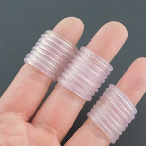 May include: Three fingers each adorned with a stack of light purple, ribbed rings. The rings are made of a translucent material, and the image is set against a dark gray background.