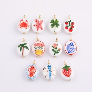 May include: A collection of white oval pendants with gold-colored loops, each hand-painted with a different design. Designs include a red crab, starfish, four-leaf clover, tomatoes, palm tree, flowers, seashell, lobster, dragonfly, and strawberry.