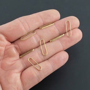 May include: A hand holding six gold oval jump rings. The jump rings are about 1/2 inch long.