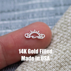 May include: A small, gold-colored charm featuring a sun and wave design. The charm is held between two fingers. Text on the image reads "14K Gold Filled Made in USA".