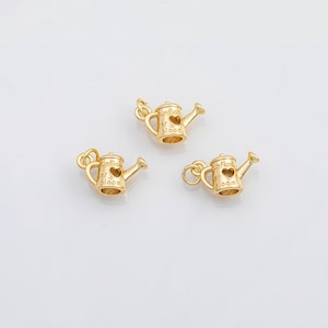 May include: Three gold-colored watering can charms. Each charm has a heart-shaped cutout and the words "unflow er bloom". The charms are small and detailed, with a loop for attaching to jewelry or other crafts.