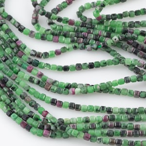 Può includere: Un filo di perline di pietre preziose verdi, nere e rosa. Le perline sono di forma quadrata e infilate su un filo trasparente.