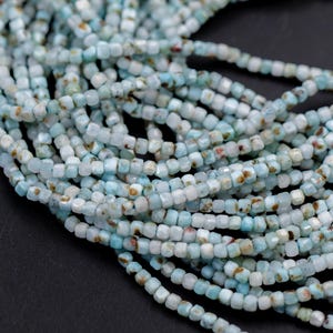 May include: A collection of light blue and white square-cut gemstone beads. The beads have a slightly irregular shape and are strung together, creating a textured surface. The beads have brown inclusions.
