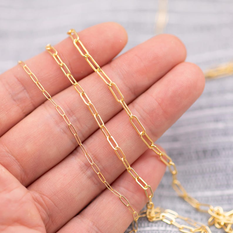 May include: Close-up of several gold-colored paperclip chain necklaces. The chains vary in size, with the links ranging from small to large. The necklaces are displayed on a light-colored surface.