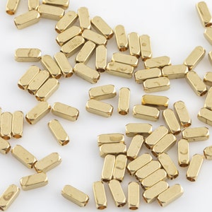 May include: A large quantity of gold-colored metal beads, each with a rectangular shape and a hole through the center. The beads are scattered on a white surface.