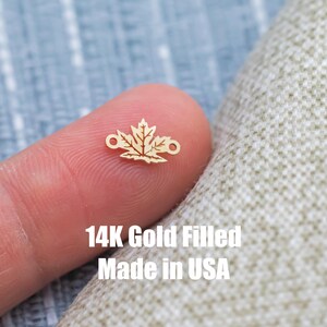May include: A small, gold-colored maple leaf charm with two small loops on either side. The charm is held between two fingers. The text "14K Gold Filled Made in USA" is visible in white.