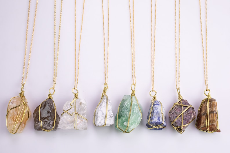 May include: A selection of gold chain necklaces, each with a distinctive, irregularly shaped gemstone pendant. The pendants display a variety of colours, including brown, clear, white, green, blue, purple, and tan. The gemstones are held in place by a gold wire cage.
