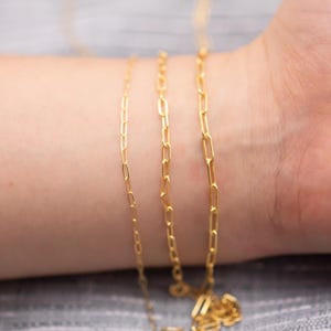 May include: Close-up of three delicate gold-colored chain necklaces. The chains have a rectangular link design. The chains are draped across a person's wrist and arm. The background is a soft gray.