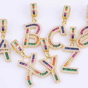 Puede incluir: Un conjunto de colgantes de oro con las letras A, B, C, Y y Z. Cada letra está incrustada con piedras preciosas de colores en forma de baguette.