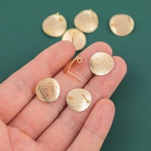 Könnte beinhalten: Goldfarbene, runde Ohrstecker mit strukturierter Oberfläche. Die Ohrringe werden auf einer Hand vor grünem Hintergrund präsentiert. Mehrere andere Ohrringkomponenten sind sichtbar.