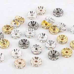 May include: A collection of round, disc-shaped metal spacer beads featuring a central hole and a row of embedded clear or iridescent rhinestones along the edge. The beads are finished in various metallic tones including gold, silver, and rose gold.