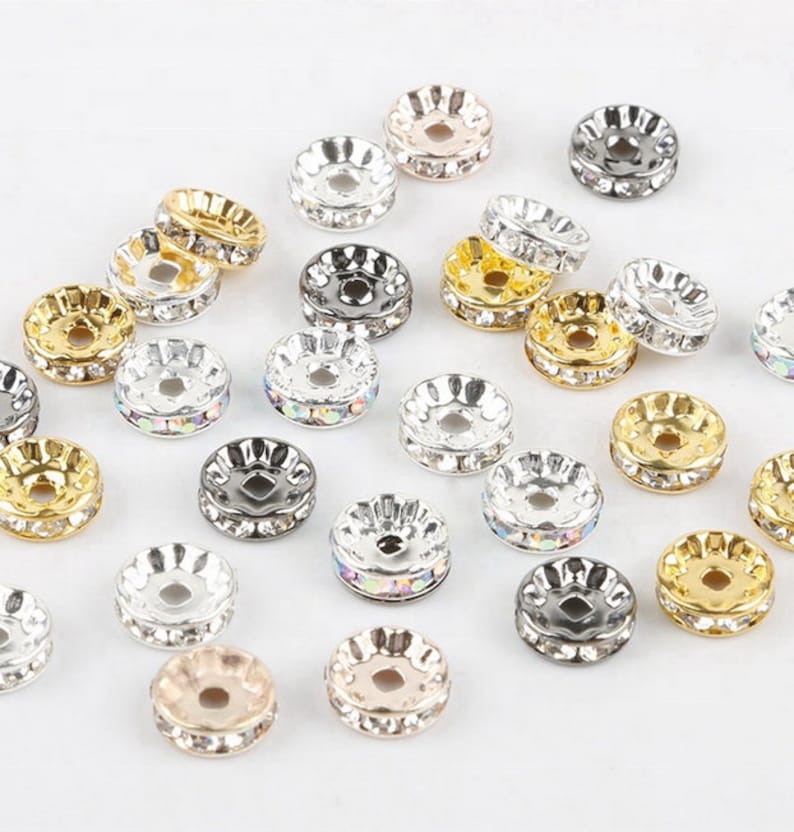 May include: A collection of round, disc-shaped metal spacer beads featuring a central hole and a row of embedded clear or iridescent rhinestones along the edge. The beads are finished in various metallic tones including gold, silver, and rose gold.