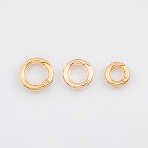 May include: Three gold-colored round spring clasps of varying sizes. The clasps are closed, with a small hinge and clasp mechanism visible. The clasps are arranged in a row against a white background, showcasing their circular design and metallic finish.