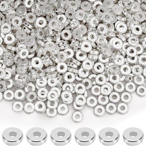 Könnte beinhalten: Eine Ansammlung glänzender, silberfarbener Metallperlen mit flacher, kreisförmiger Form und einem Loch in der Mitte. Die Perlen sind verstreut, einige sind in einer Reihe am unteren Rand angeordnet. Geeignet für Schmuckherstellung oder andere Bastelarbeiten.