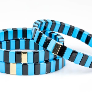 May include: Three stacked bracelets made of black and light blue ceramic tiles. The bracelets have gold accents.