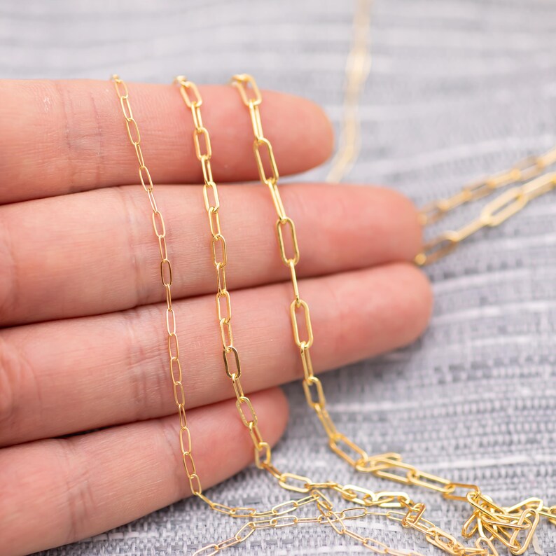 May include: Close-up of three delicate gold-colored chain necklaces with a paperclip link design. The chains are displayed against a neutral gray background, showcasing their fine details and polished finish. The chains are held in a hand.