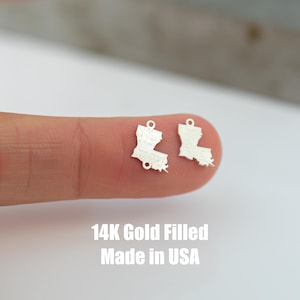 Peut inclure: Deux breloques en or 14 carats remplies en forme de l'État de la Louisiane. Les breloques sont sur un doigt et le texte "14K Gold Filled Made in USA" est sous les breloques.
