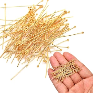 May include: A collection of gold-colored head pins, scattered across a white surface. Each pin features a slender wire shaft topped with a small, round bead. A small bundle of pins is held in a hand, showcasing the product's quantity and detail.