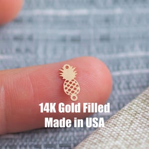 Puede incluir: Un pequeño dije de piña de color dorado, con una superficie texturizada. El dije tiene un lazo en la parte inferior y una corona detallada. El texto "14K Gold Filled Made in USA" es visible debajo.