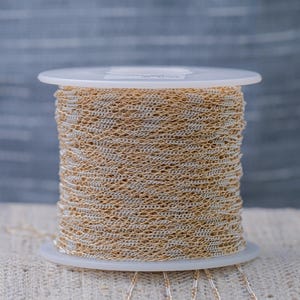 May include: A spool of gold and silver-colored chain. The chain is tightly wound around a white plastic spool. The background is a blurred gray and white. The chain is likely used for jewelry making or other crafts.