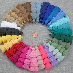 May include: A collection of colourful tassel trims in various shades of blue, green, yellow, red, pink, and brown. The tassels are arranged in a circle on a grey textured background.