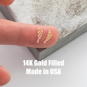 Op de afbeelding: Twee kleine, goudkleurige vleugelbedels met gedetailleerde veermotieven. De bedels worden op een vinger getoond, met de witte tekst "14K Gold Filled Made in USA".