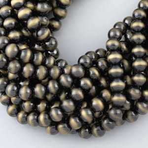 May include: A collection of round beads arranged in a curved formation. Each bead features a metallic finish with a blend of dark charcoal grey and muted gold tones, creating a reflective surface with a distinct horizontal band of lighter colour.