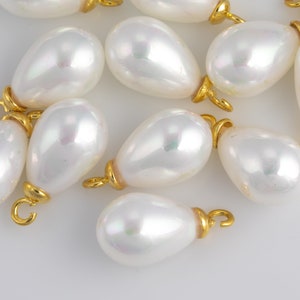 May include: A group of white pearlescent teardrop-shaped beads with gold-toned metal loops. The beads are arranged on a white surface.