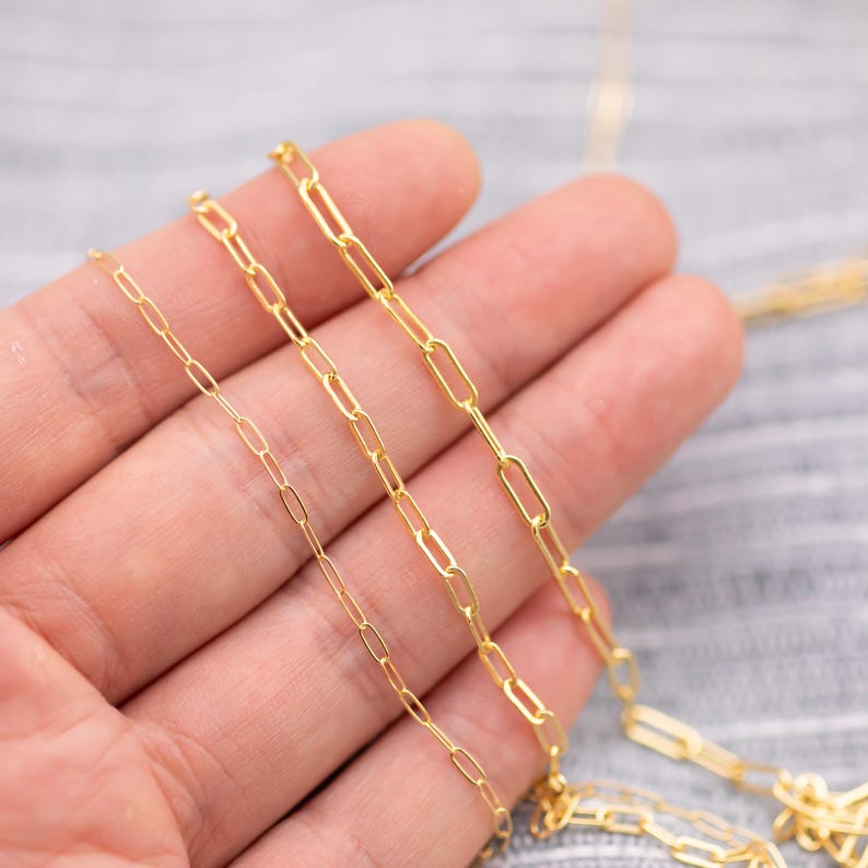 May include: Close-up of three gold-colored chain necklaces with a paperclip link design. The chains are displayed on a light-colored surface. The chains vary in link size, with the smallest chain on the left.
