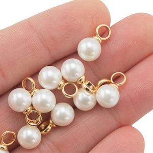 May include: A close-up of a hand holding a group of white pearl charms with gold jump rings. The charms are perfect for adding a touch of elegance to jewelry projects.
