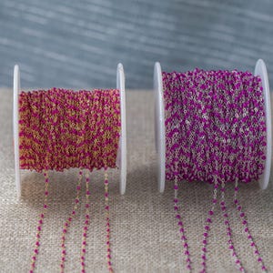 Puede incluir: Dos carretes de cadena de cuentas. Uno presenta una cadena dorada con cuentas rosas, el otro una cadena plateada con cuentas rosas. Los carretes blancos están sobre una superficie neutra, ideales para bisutería.