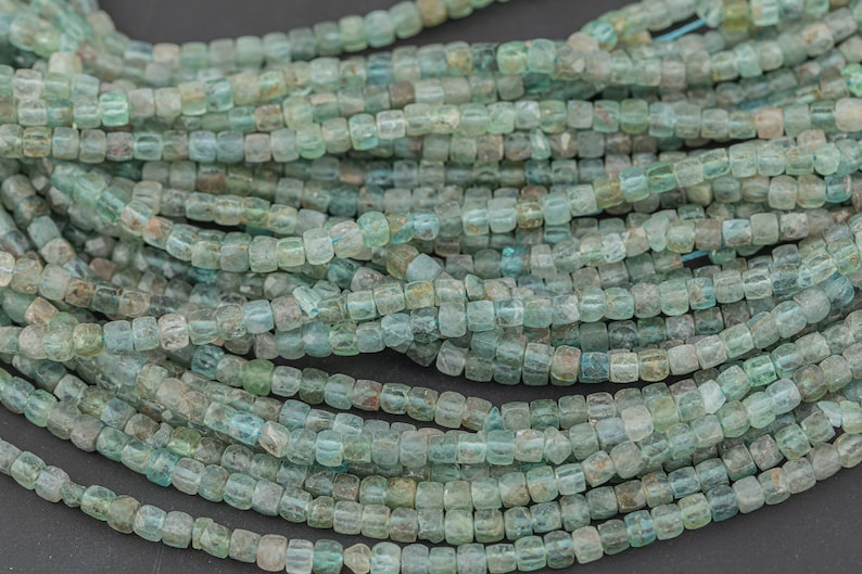 May include: Strands of small, square-shaped beads in shades of teal, turquoise, and muted green. The beads are strung together, creating a textured and layered effect. Ideal for jewelry making or crafting projects.