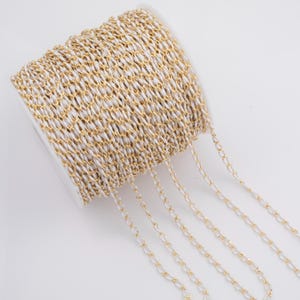 May include: A spool of gold-colored chain with a white woven backing. The chain is made of small, oval-shaped links. The spool is on a white background.
